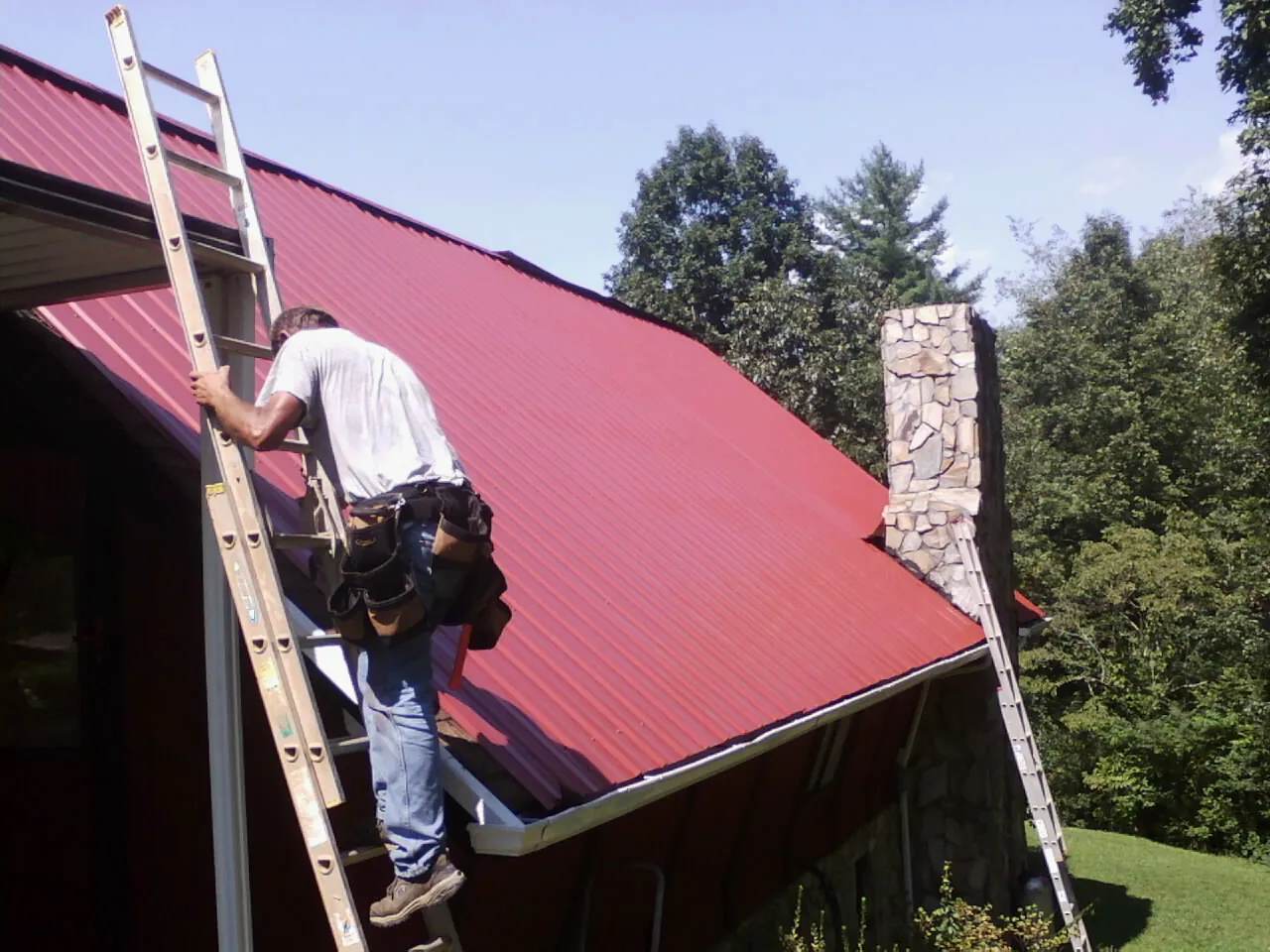 Roofer on a ladder beside a red metal roof and stone chimney — Soffit & Fascia Repair by Roofing Brothers in Huntington Park